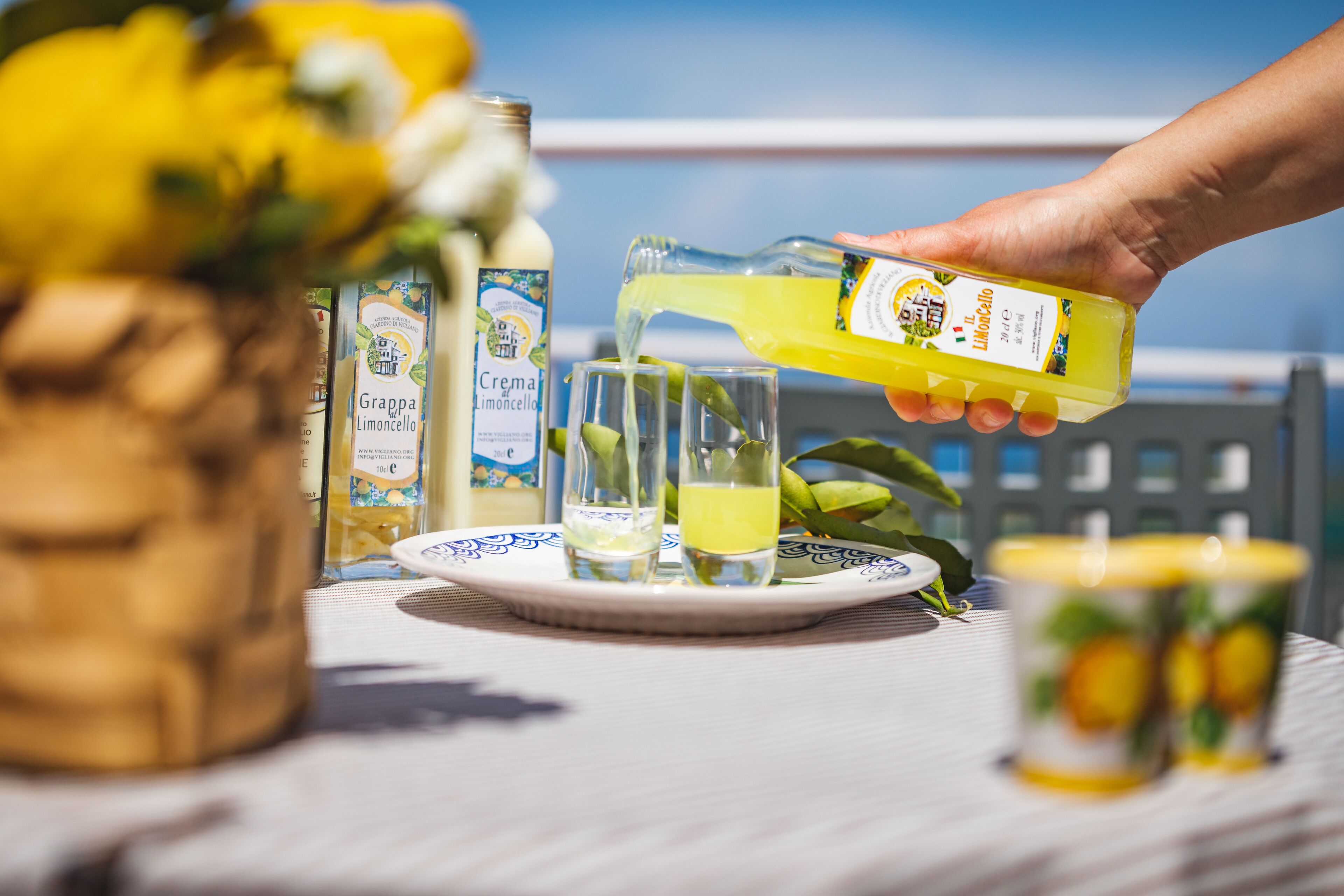 Une main verse une boisson jaune dans des verres sur une table, à côté d’une assiette blanche et de bouteilles de Crema Limoncello.