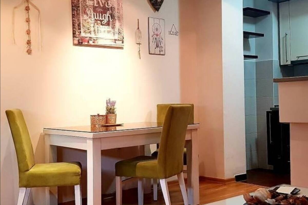 A dining area with a white table and two green chairs, a decorative wall hanging, and a kitchen area with cabinets.