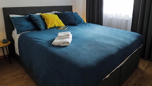 A neatly made bed with a dark blue bedspread and pillows, a small bedside table with a lamp, and a window with sheer curtains. - Ana 2 (Banja Luka)