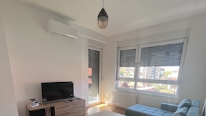 A modern living room with a wooden floor, a grey sofa, a coffee table, a TV on a cabinet, and a window with blinds. - Ana 2 (Banja Luka)