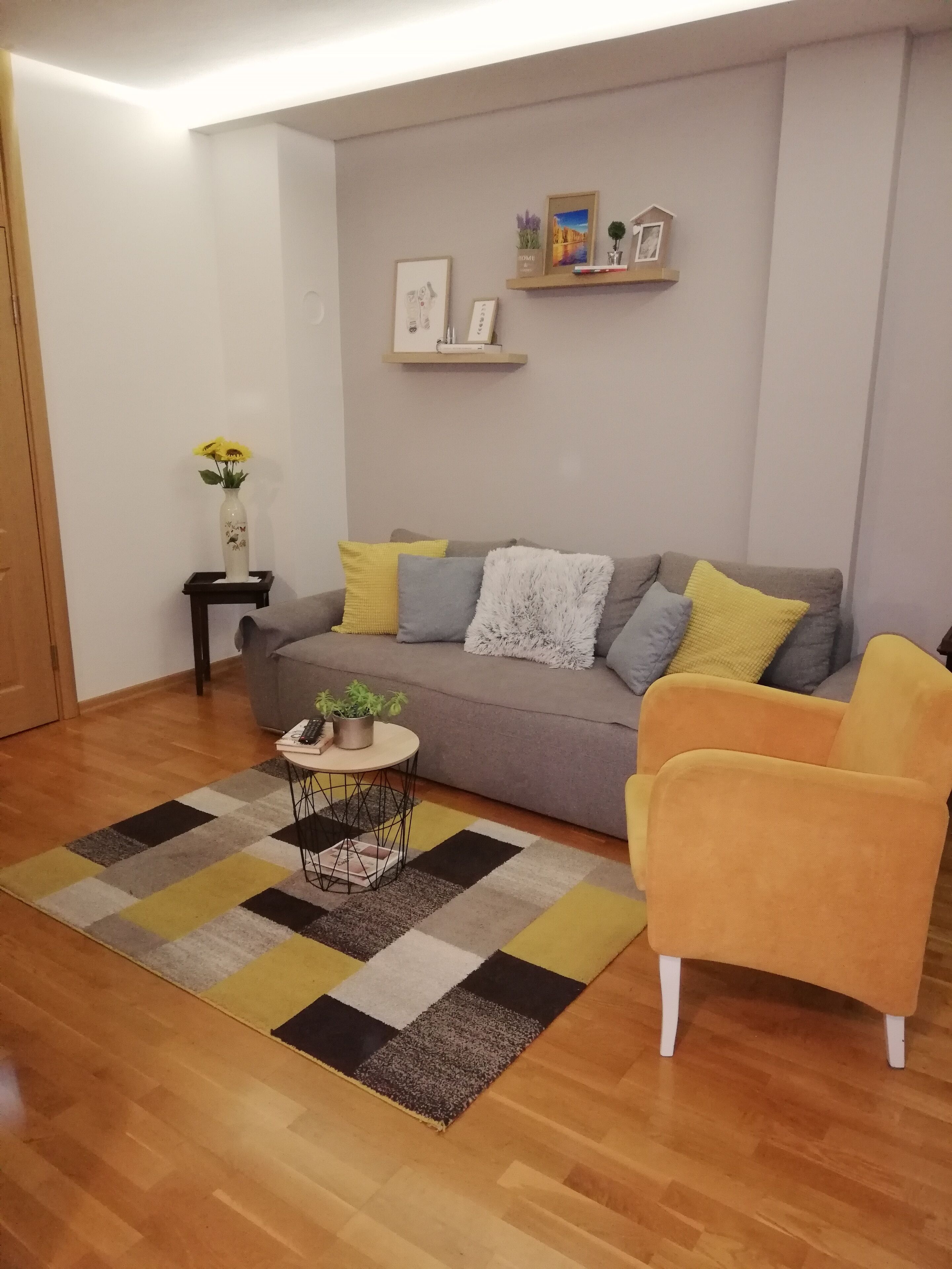 Uma sala de estar com um sofá cinza, almofadas amarelas e cinzas, uma poltrona amarela, uma pequena mesa redonda com uma planta e um tapete xadrez.