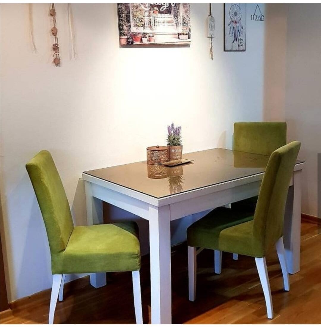 A dining table with four green upholstered chairs, a glass top, and a small plant on the table.