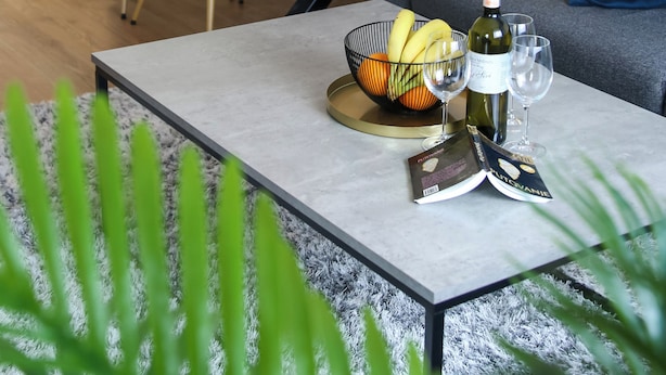 A modern living room with a dining area, featuring a sofa with blue and yellow cushions, a coffee table with a wine bottle and glasses, and a dining table set with plates and a vase of red flowers. - Ana 2 (Banja Luka)