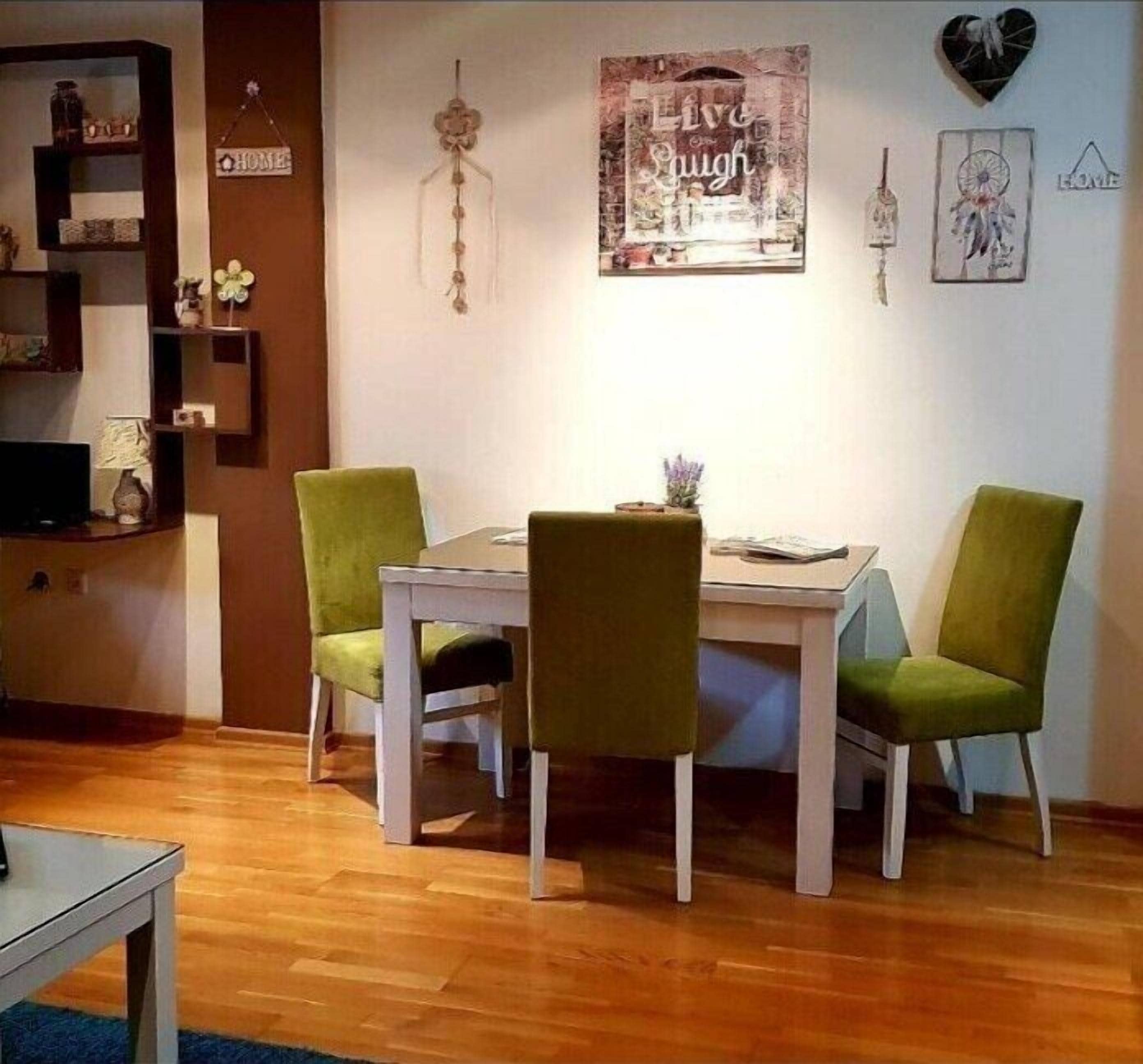 Un coin repas avec une table blanche et des chaises vertes, un mur orné d’une inscription « Vivez, aimez, riez », et une bibliothèque contenant divers objets.