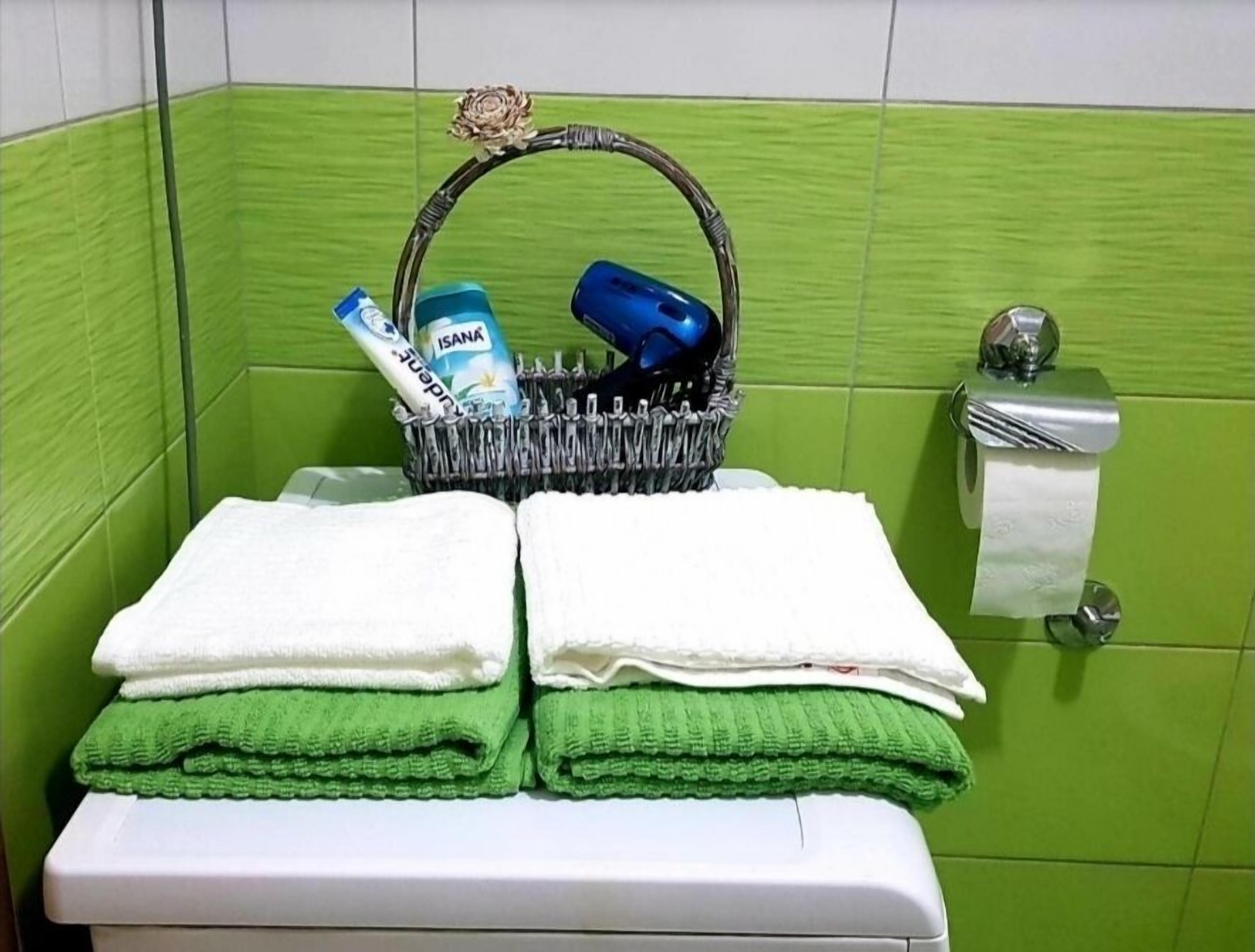 A bathroom with green tiles, a white sink, a silver faucet, a white toilet, and a basket containing toiletries.