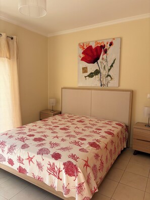 A bedroom with a bed, a nightstand, a lamp, a painting, and a window with curtains.