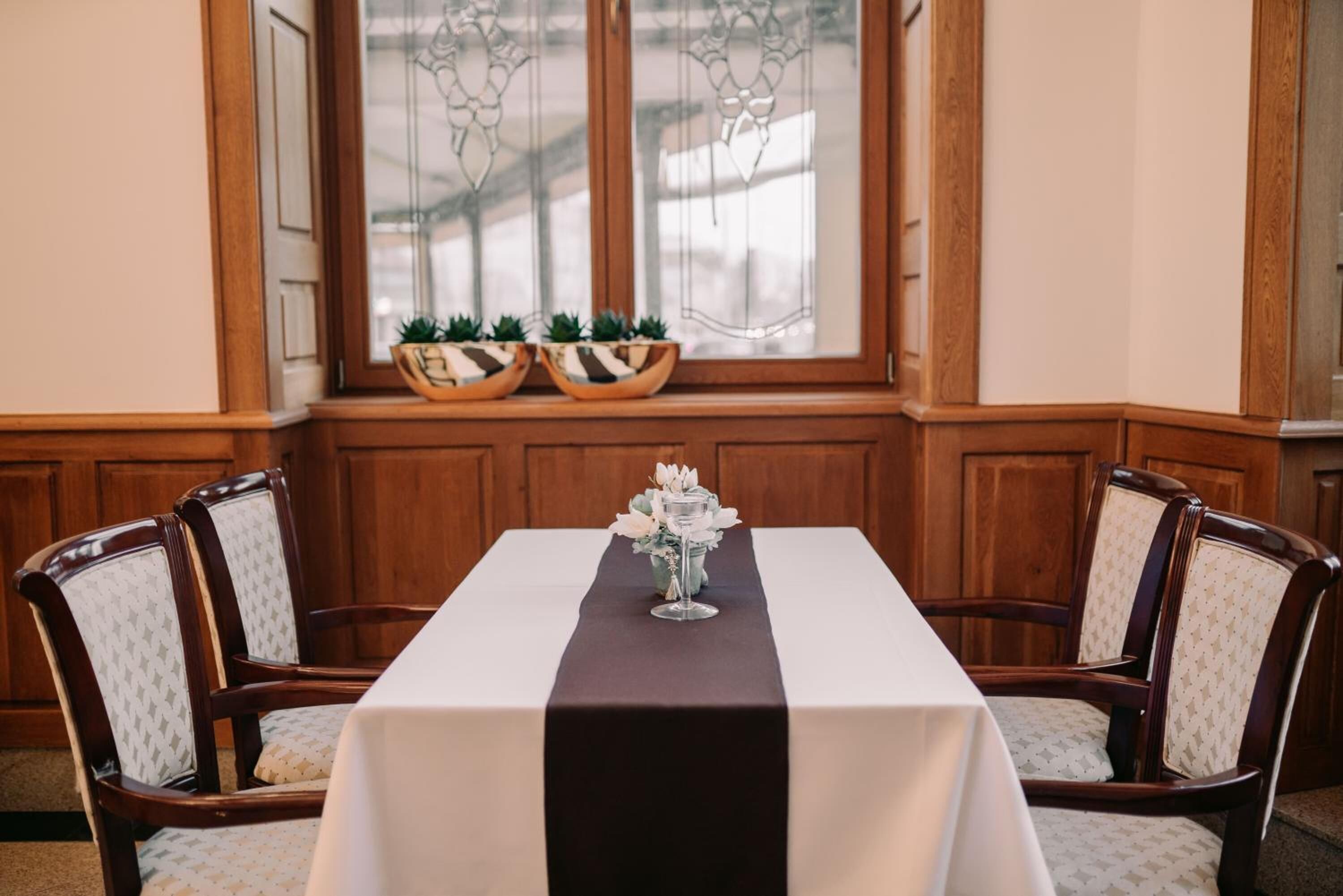 A dining table with a black runner, surrounded by four upholstered chairs with a star pattern. There are two potted plants on the windowsill above the table.