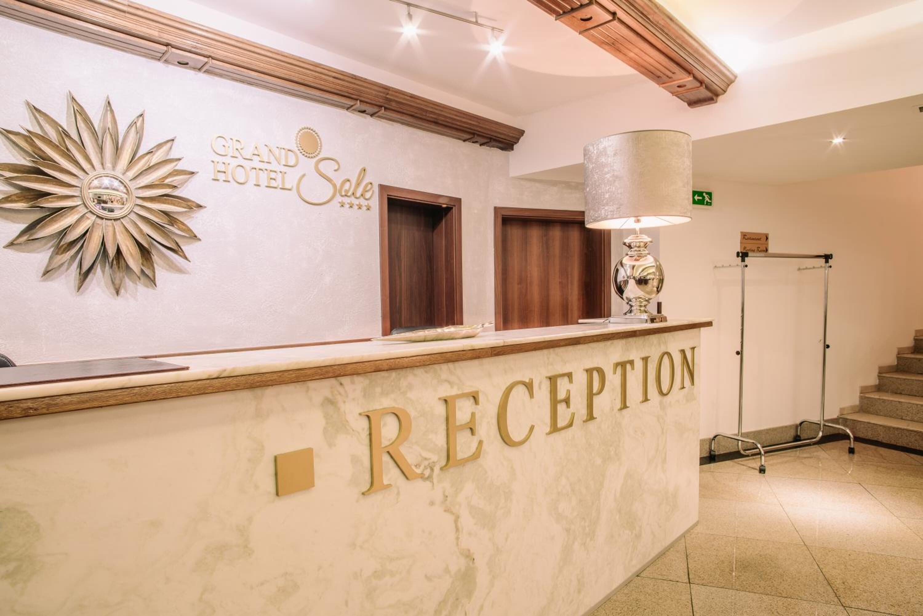 Uma área de recepção de hotel com um balcão de mármore, uma grande decoração em formato de sol e uma placa com a inscrição 'Grand Hotel Sale'.