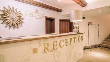 Uma área de recepção de hotel com um balcão de mármore, uma grande decoração em formato de sol e uma placa com a inscrição 'Grand Hotel Sale'.