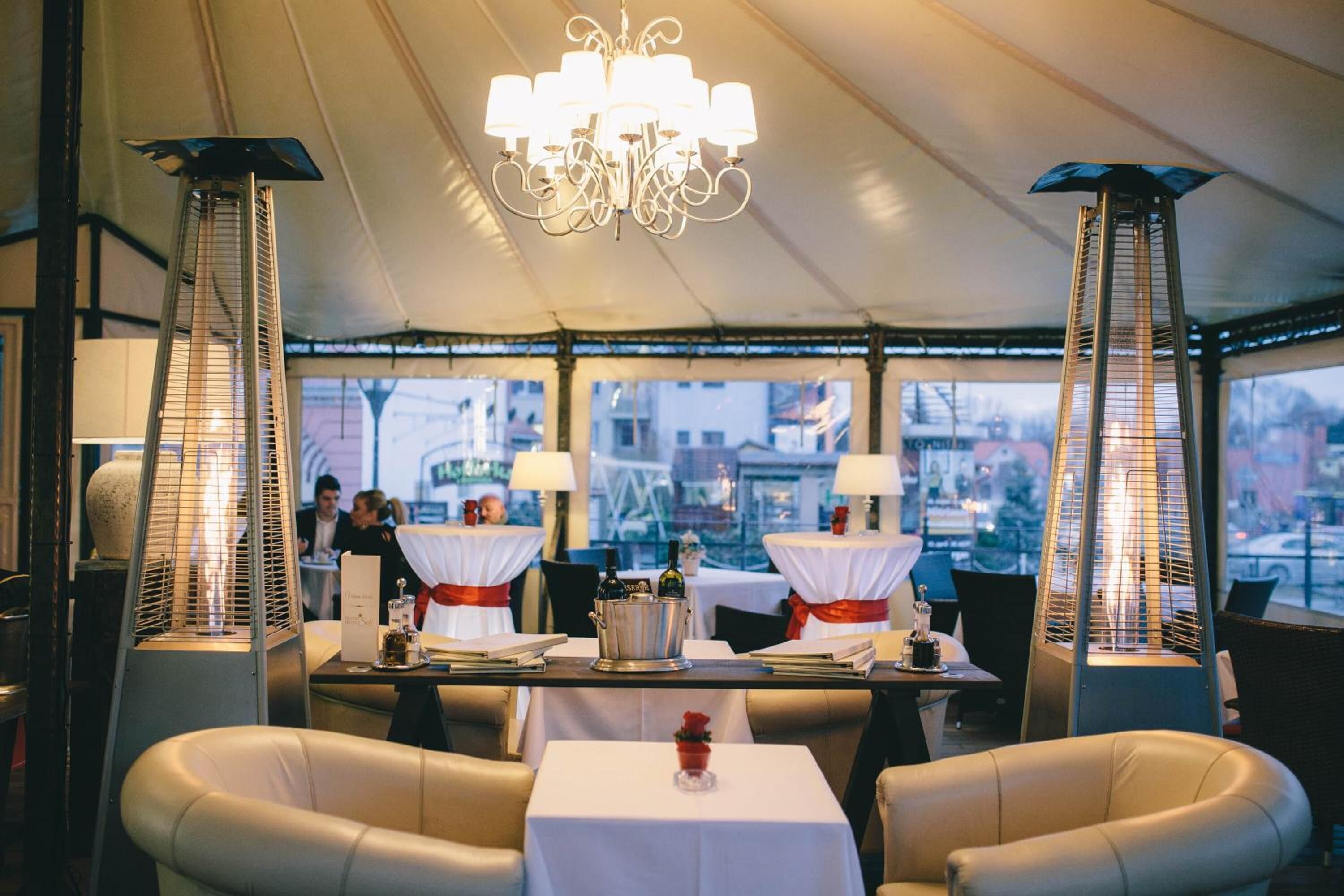 En restaurang med dukade bord, en stor kristallkrona och stora fönster med utsikt över staden.
