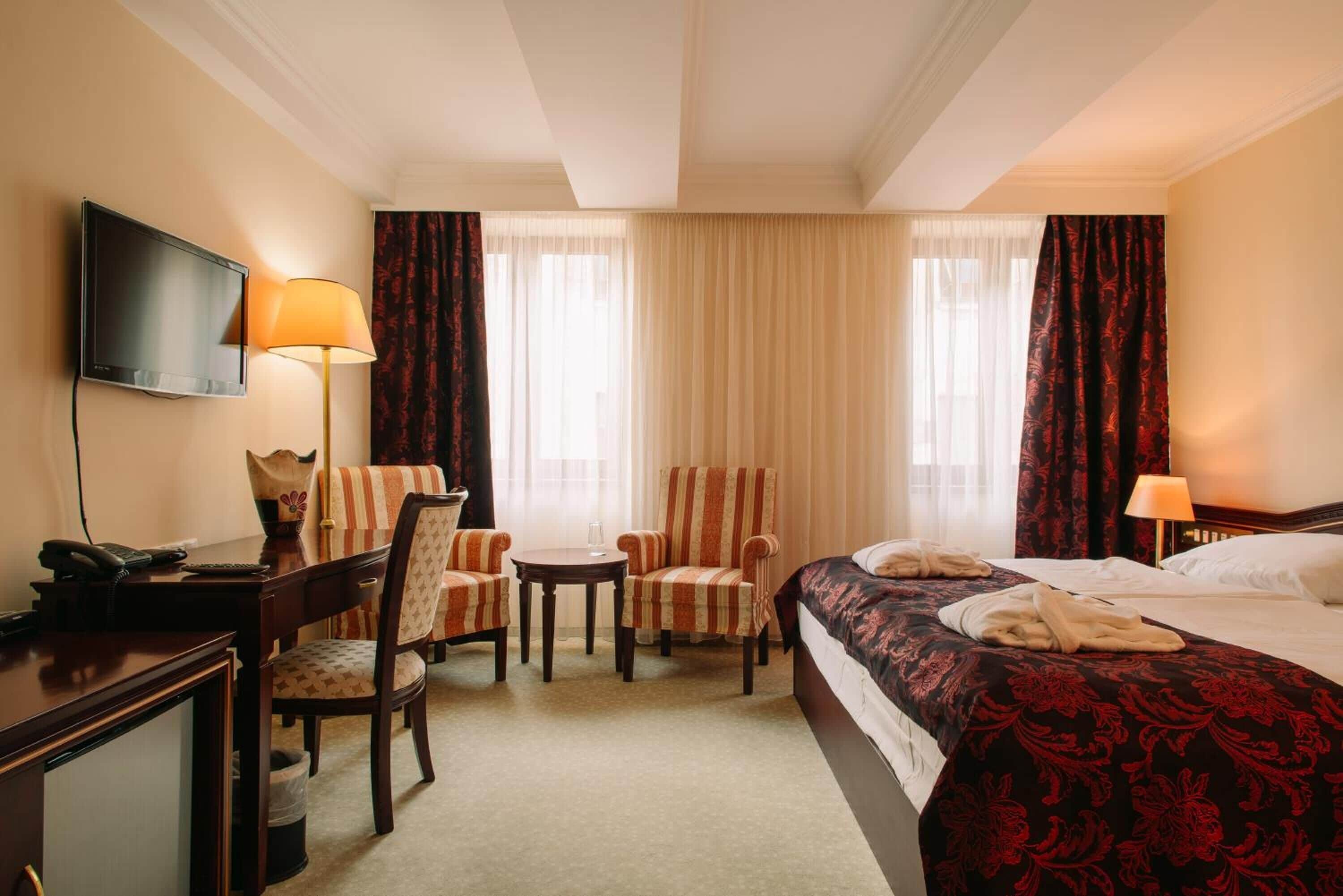 A hotel room with a bed, two armchairs, a small table, a desk with a telephone, and a television.