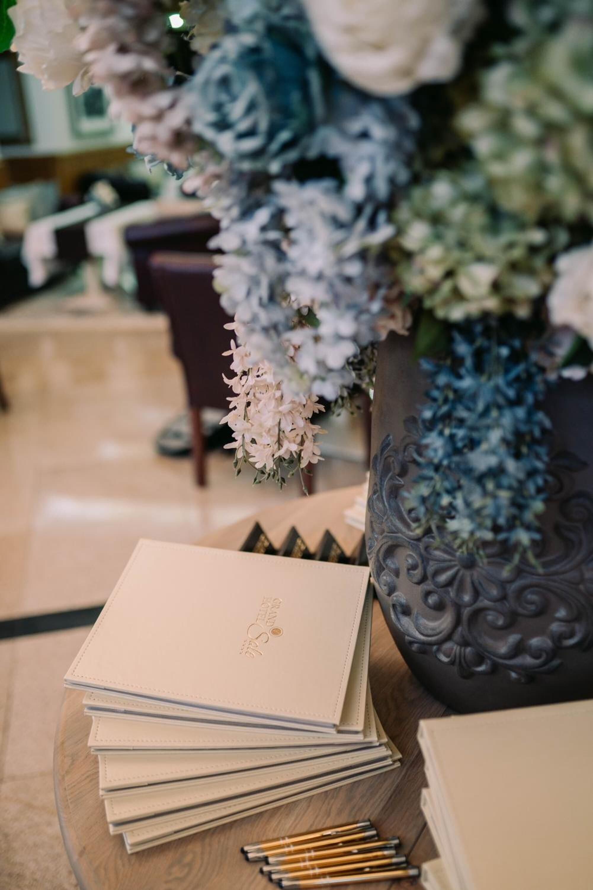 A stack of notebooks on a table with a vase of flowers in the background.