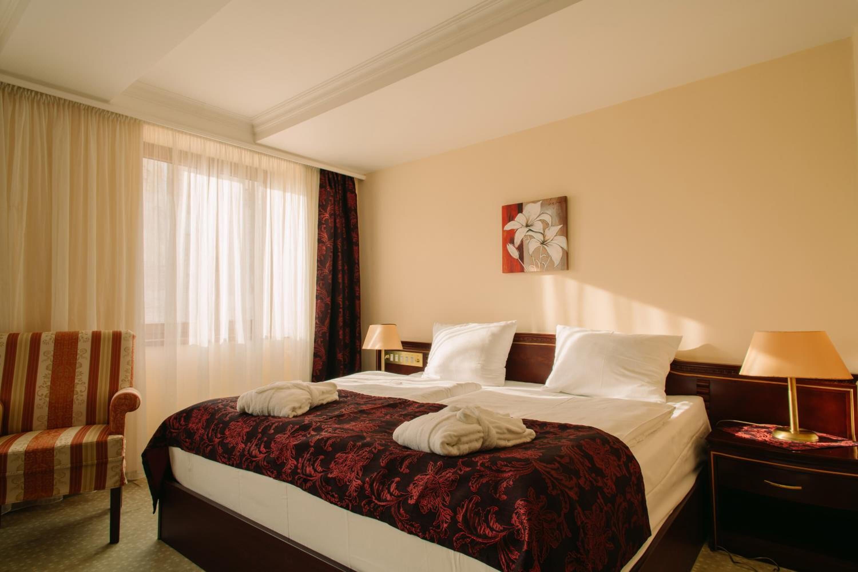 A hotel room with a large bed, two bedside tables with lamps, a chair, and a window with sheer curtains.
