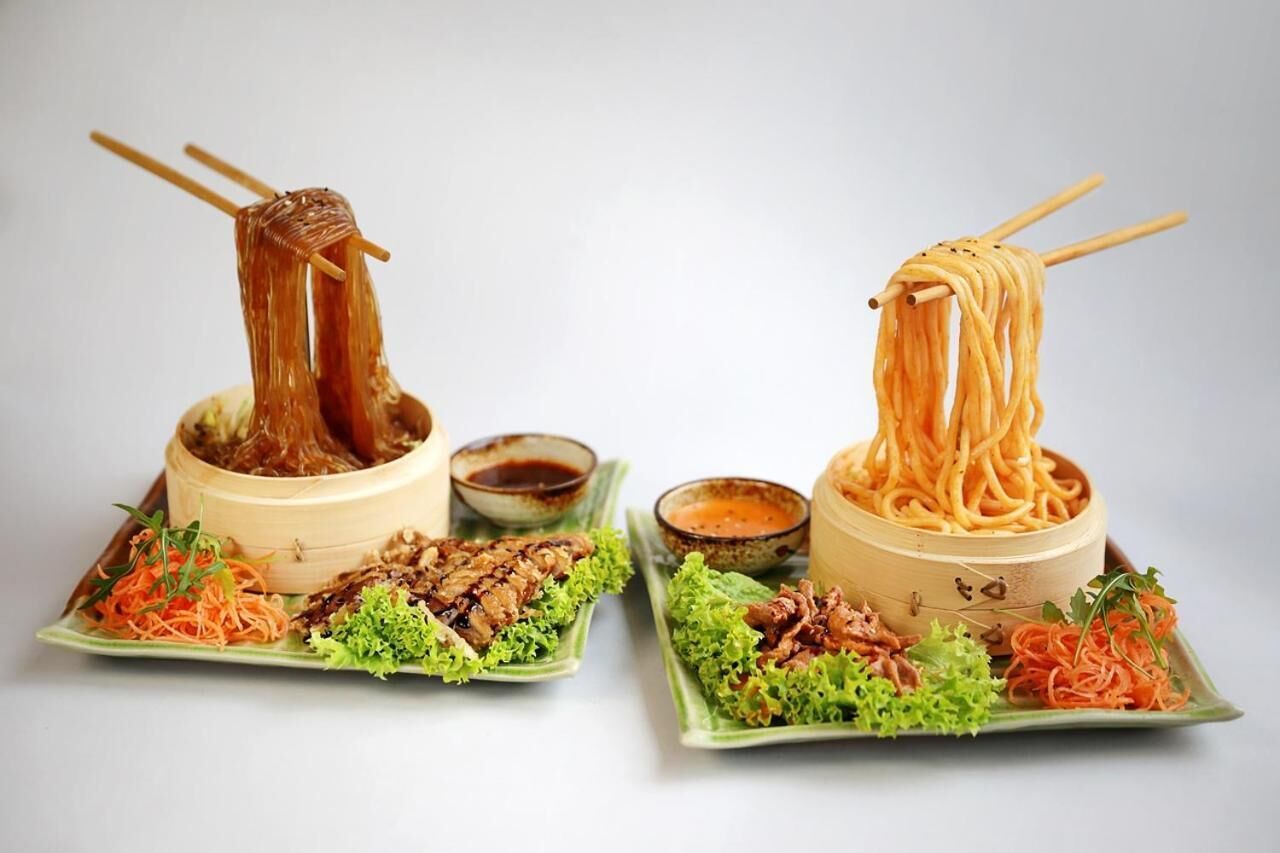 Two bowls of noodles with chopsticks, accompanied by small bowls of sauce and garnishes.