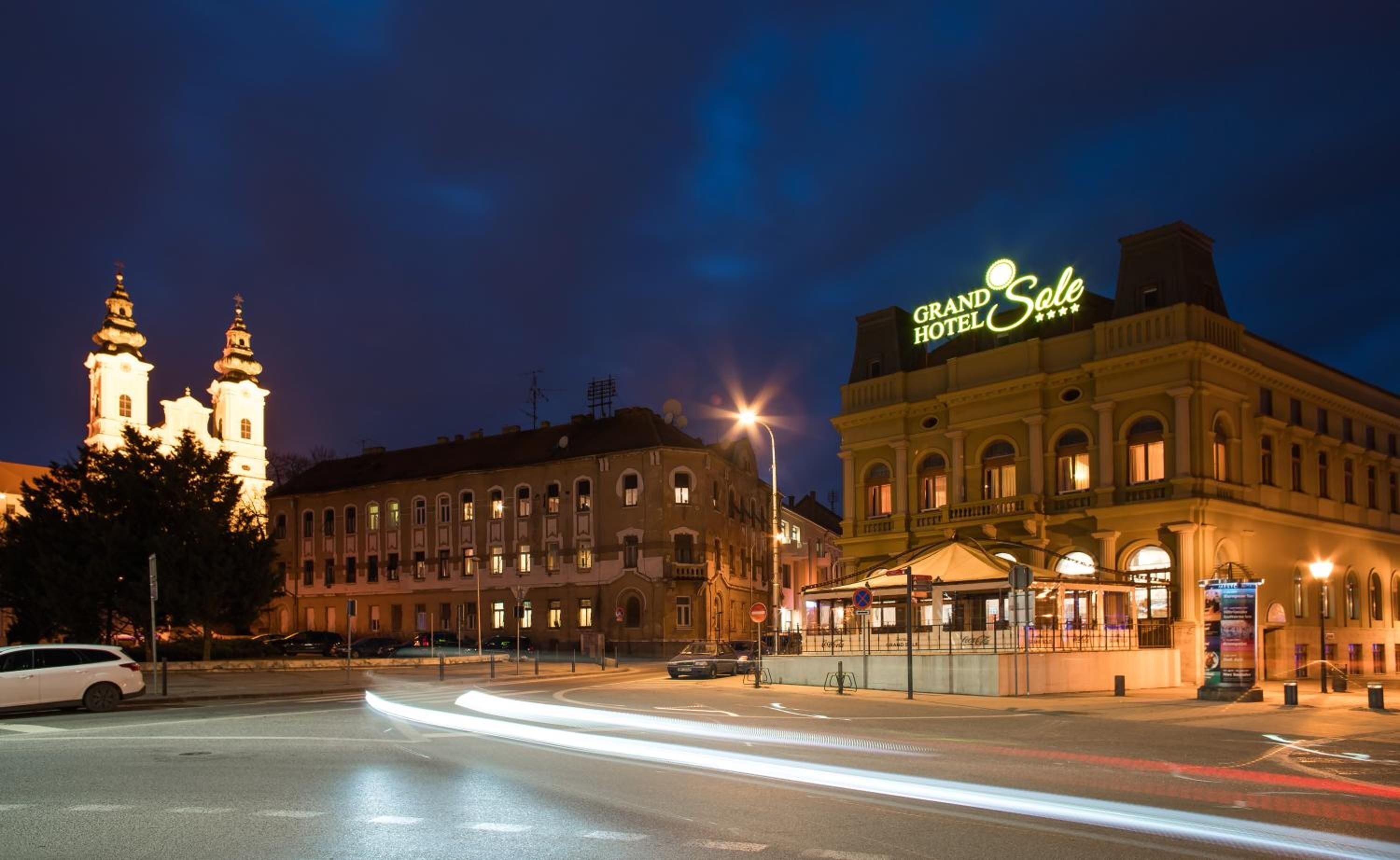 Hotel Grand Sorel con un letrero llamativo, un edificio histórico con dos torres al fondo y una calle bien iluminada en primer plano.