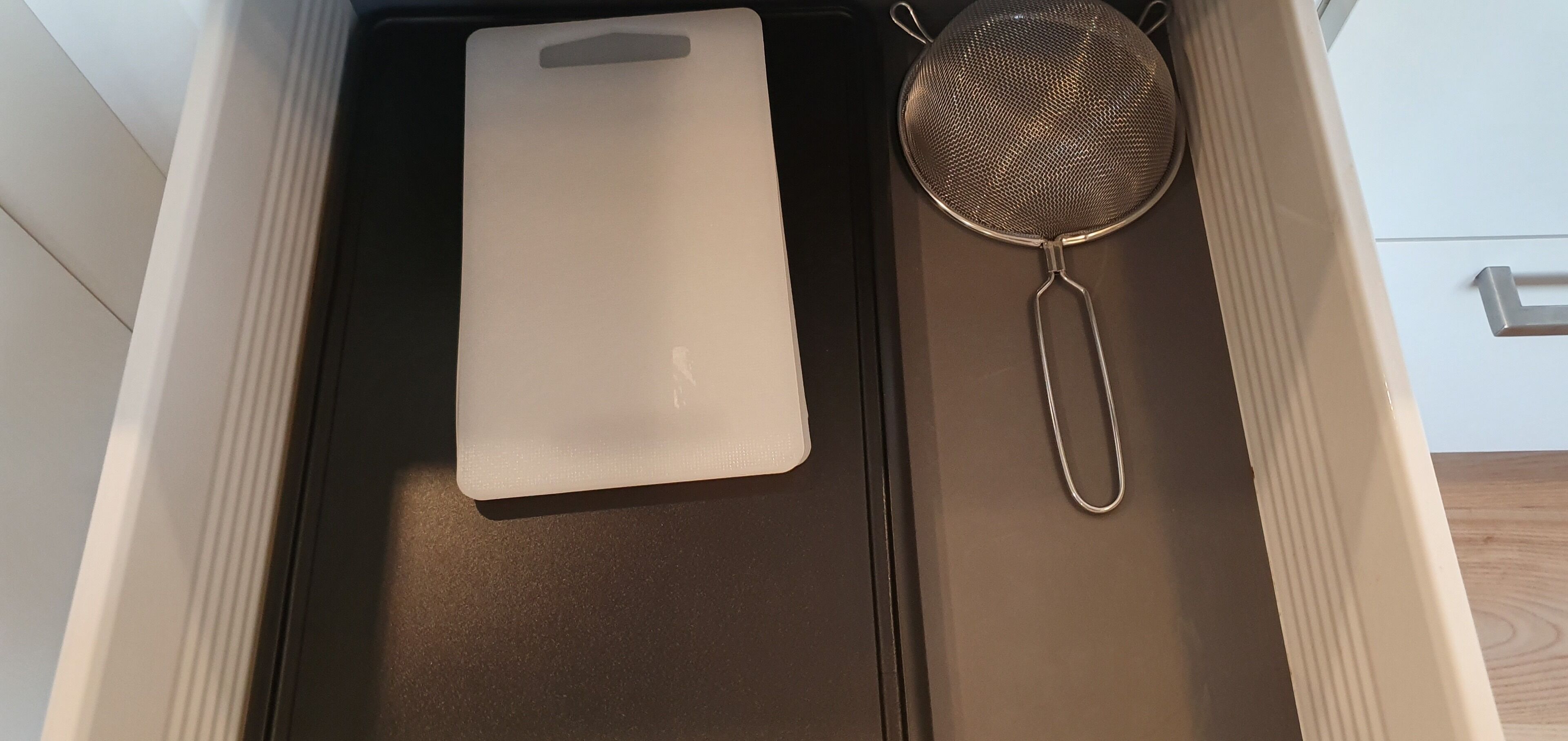 Uma gaveta de cozinha com uma tábua de corte branca e um escorredor de metal.