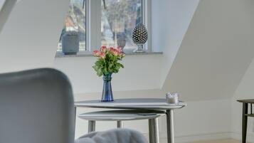 Una habitación con una pequeña mesa, una silla, un florero con flores y una ventana con un adorno.