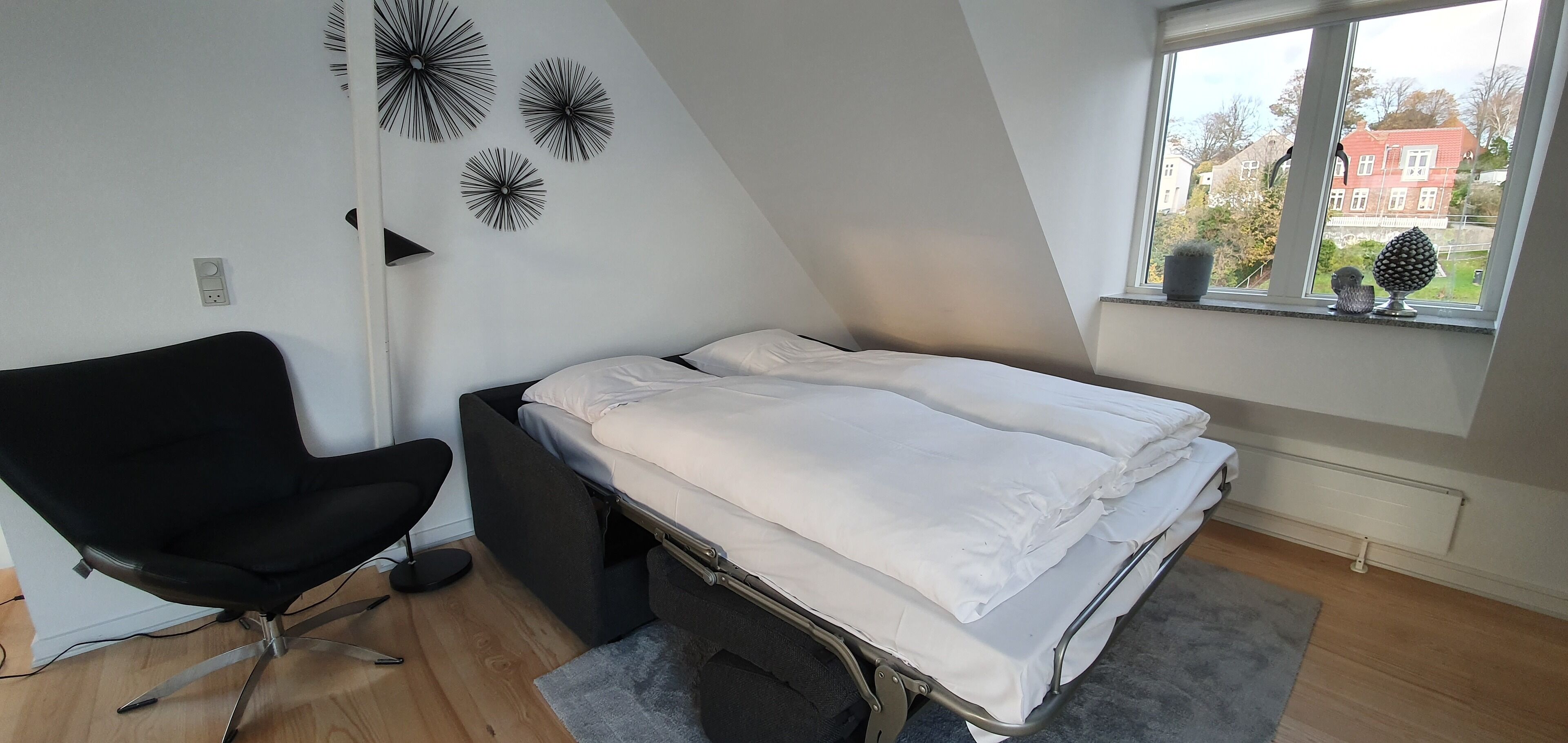 Ett litet, modernt sovrum med en säng, en svart stol och ett fönster med utsikt över hus.