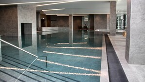 En rummelig indendørs swimmingpool med klart vand, omgivet af stenvægge og moderne lysarmaturer.