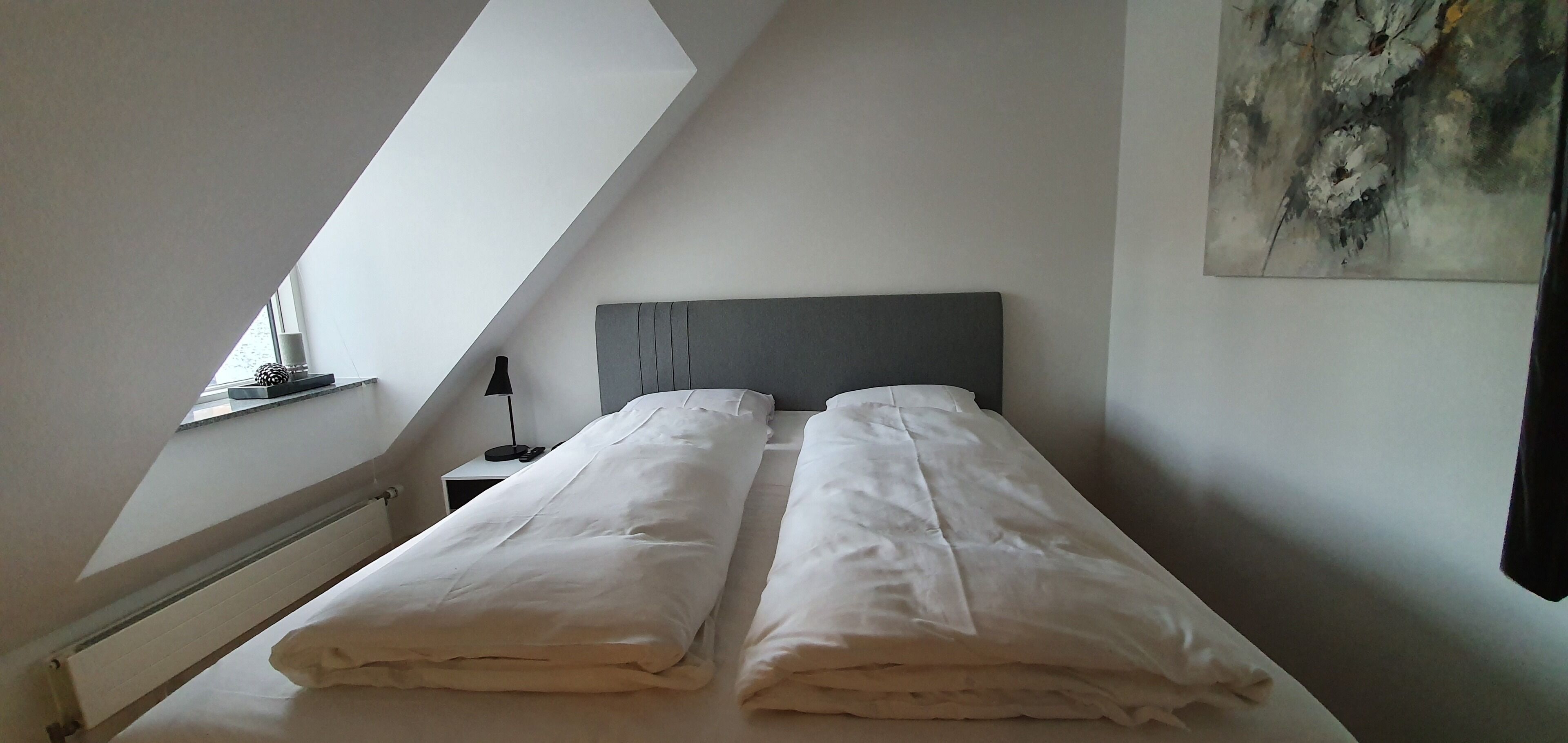 Een slaapkamer met schuine plafonds, een bed met witte lakens, een nachtkastje met een lamp en een schilderij aan de muur.