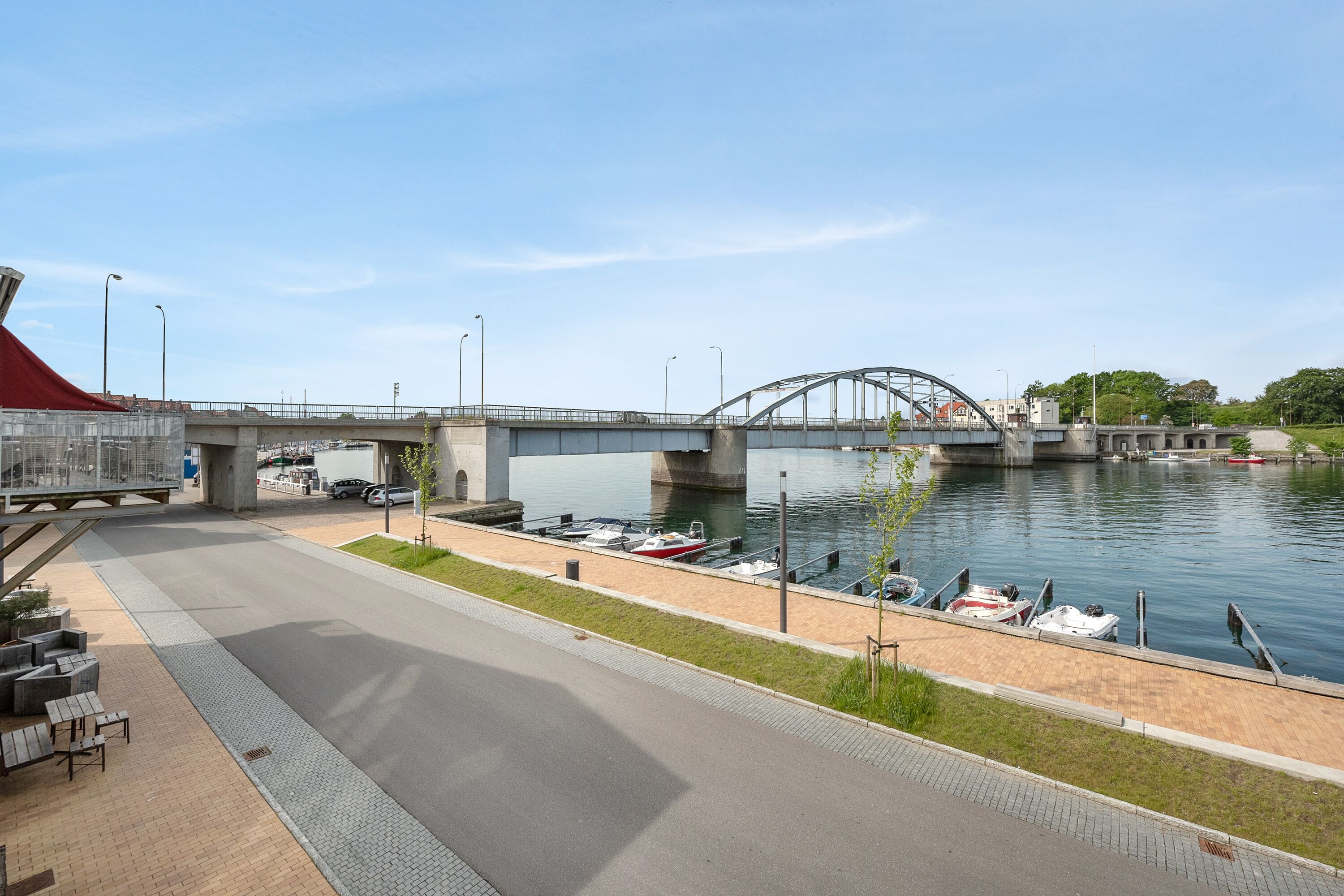 A riverside walkway with a bridge, boats docked, and a red tent-like structure.
