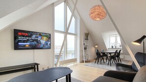En moderne stue med et tv på væggen, et spiseområde med bord og stole, og et stort vindue der giver naturligt lys.