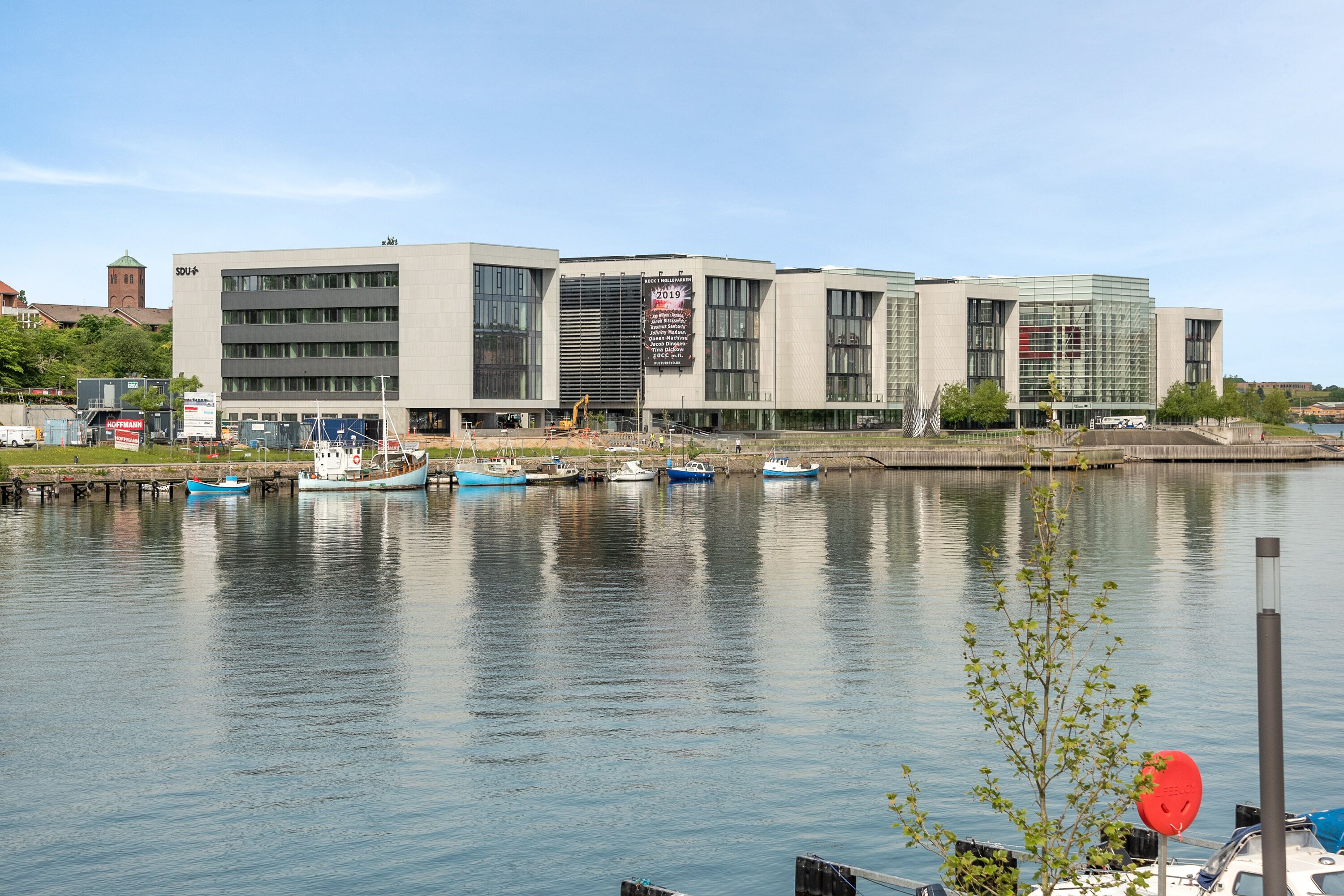 A modern building with multiple floors and large windows, situated next to a calm body of water with several boats docked.