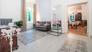 A spacious living room with a grey sofa, a glass coffee table, and a chandelier. There is a dining area with a wooden table and chairs visible through an open doorway.