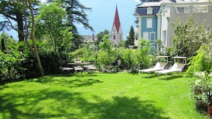 A lush green lawn with lounge chairs and a dining table, surrounded by trees and a building with a red roof in the background.