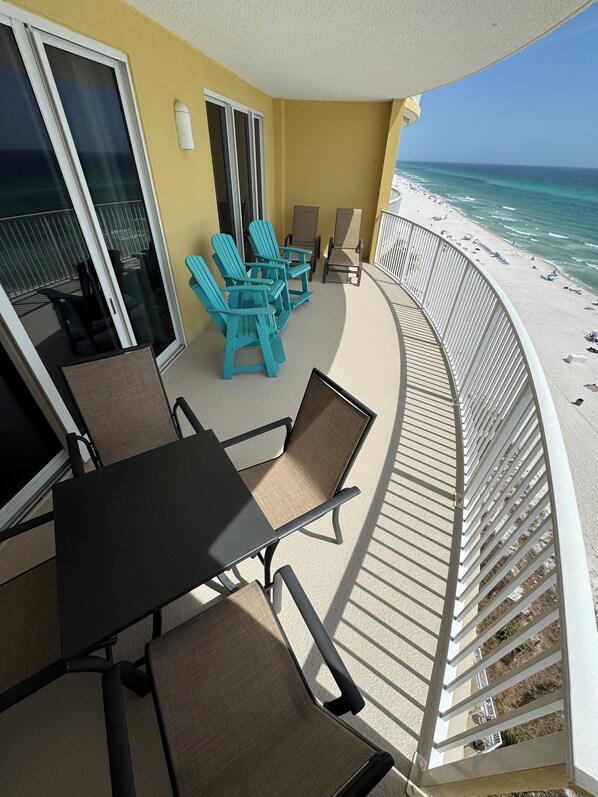 A balcony with a table and chairs, a view of the beach, and a glass door leading to a shaded area.
