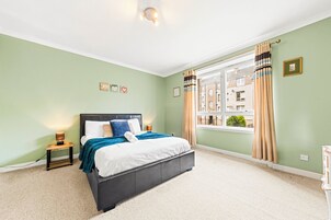 Un dormitorio con cama, mesitas de noche, una ventana con cortinas y vistas a edificios.