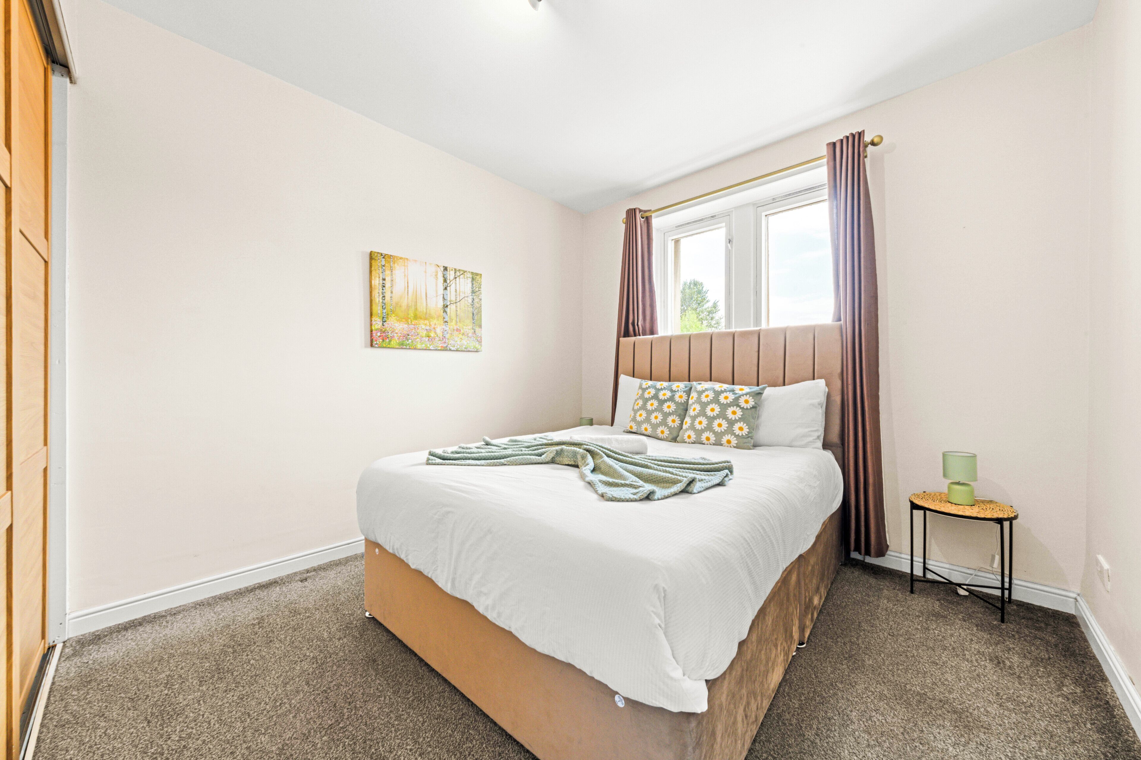 Een slaapkamer met een bed, een raam met gordijnen, een klein tafeltje met een lamp en een schilderij aan de muur.