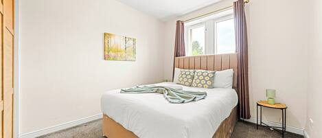 Een slaapkamer met een bed, een raam met gordijnen, een klein tafeltje met een lamp en een schilderij aan de muur.