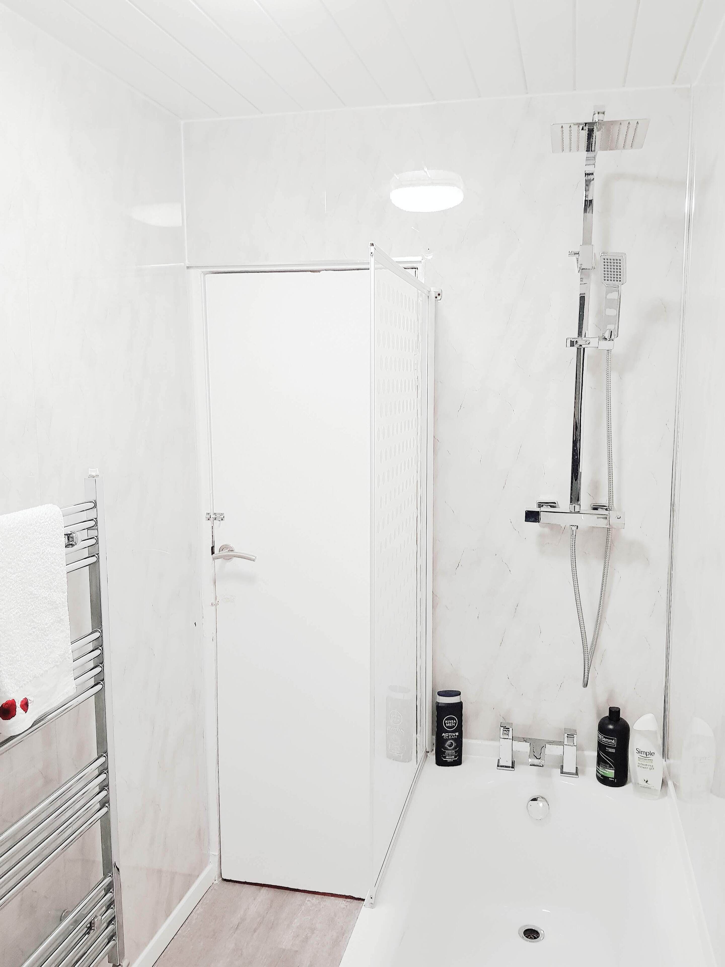A bathroom with a white door, a shower with a handheld showerhead, a towel rack with towels, and toiletries on the sink.