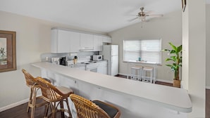 A modern kitchen with white cabinets, a central island, bar stools, a ceiling fan, and a window with blinds. - Beachfront Hideaway in the heart of Key Largo! Your own private beach! (Key Largo)