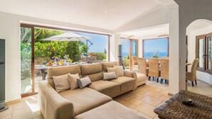 A modern living room with a beige sofa, wicker chairs, a dining table with outdoor seating, and a view of the sea.