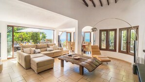 A spacious living area with a sofa set, a coffee table, and a dining table. There are large windows with views of greenery and a balcony.
