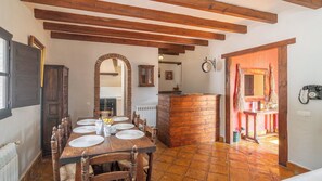 Un coin repas rustique avec une table en bois dressée pour quatre personnes, une arche en briques et une cuisine visible au fond.
