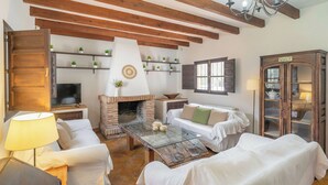 Ein gemütliches Wohnzimmer mit Kamin aus Ziegelsteinen, weißen Sofas, einem Glastisch, Holzbalken an der Decke und einer Schranke mit Glastüren.