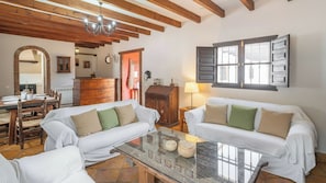 Un salon chaleureux avec deux canapés blancs, une table basse en verre, des poutres en bois au plafond, un coin repas avec une table et des chaises en bois, et une grande fenêtre avec des volets.