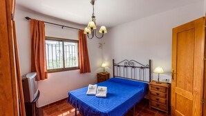 A bedroom with a bed, two bedside tables, a wooden wardrobe, a window with curtains, a television, and a chandelier. - Casa Los Pérez Sedella by Ruralidays (Málaga)