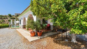 A building with a white facade, arched doorways, and a gravel driveway. There are potted plants and a large tree in front of the building. - Casa Los Pérez Sedella by Ruralidays (Málaga)