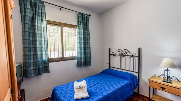 Un lit simple avec une couvre-lit bleue, une table de chevet en bois avec une lampe, et une fenêtre avec des rideaux verts.