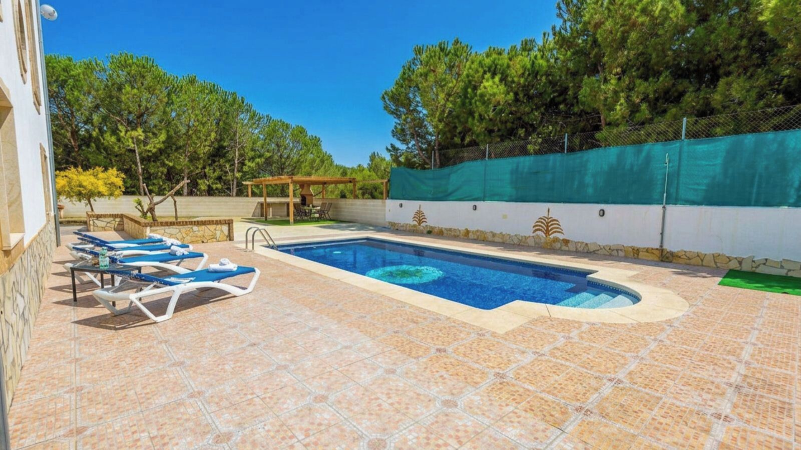 Piscina con sillones de descanso y una pérgola, rodeada de un patio con suelo de piedra y una cerca verde.