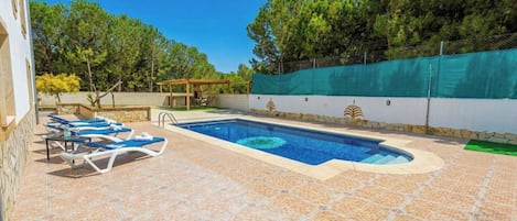 Piscina con sillones de descanso y una pérgola, rodeada de un patio con suelo de piedra y una cerca verde.