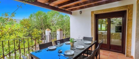 Un área de comedor al aire libre con una mesa dispuesta para cuatro personas, rodeada de sillas de mimbre, bajo una pérgola de madera.