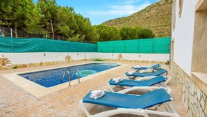 Une piscine dotée d’une terrasse carrelée, entourée de chaises longues avec des serviettes, et un mur de pierre surmonté d’un treillis vert.