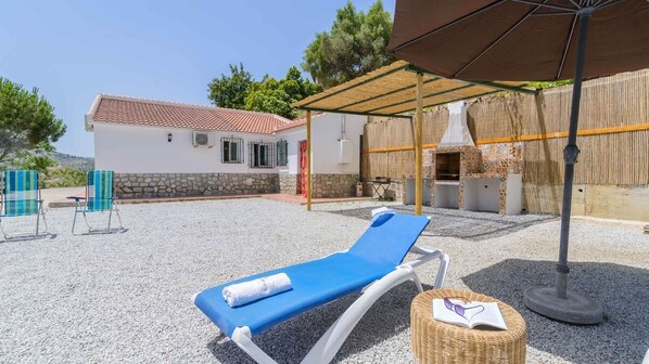 A covered outdoor area with a lounge chair, a wicker basket with sunglasses, and a grill area.