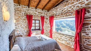 Un dormitorio con paredes de piedra, una cama, cortinas rojas, una ventana con vistas a la montaña y un techo de madera.