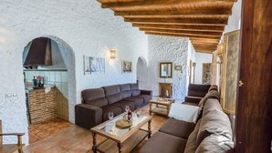 Un interior rústico con pared de piedra, vigas de madera, un sofá, una mesa de centro y una zona de cocina con fregadero y utensilios.